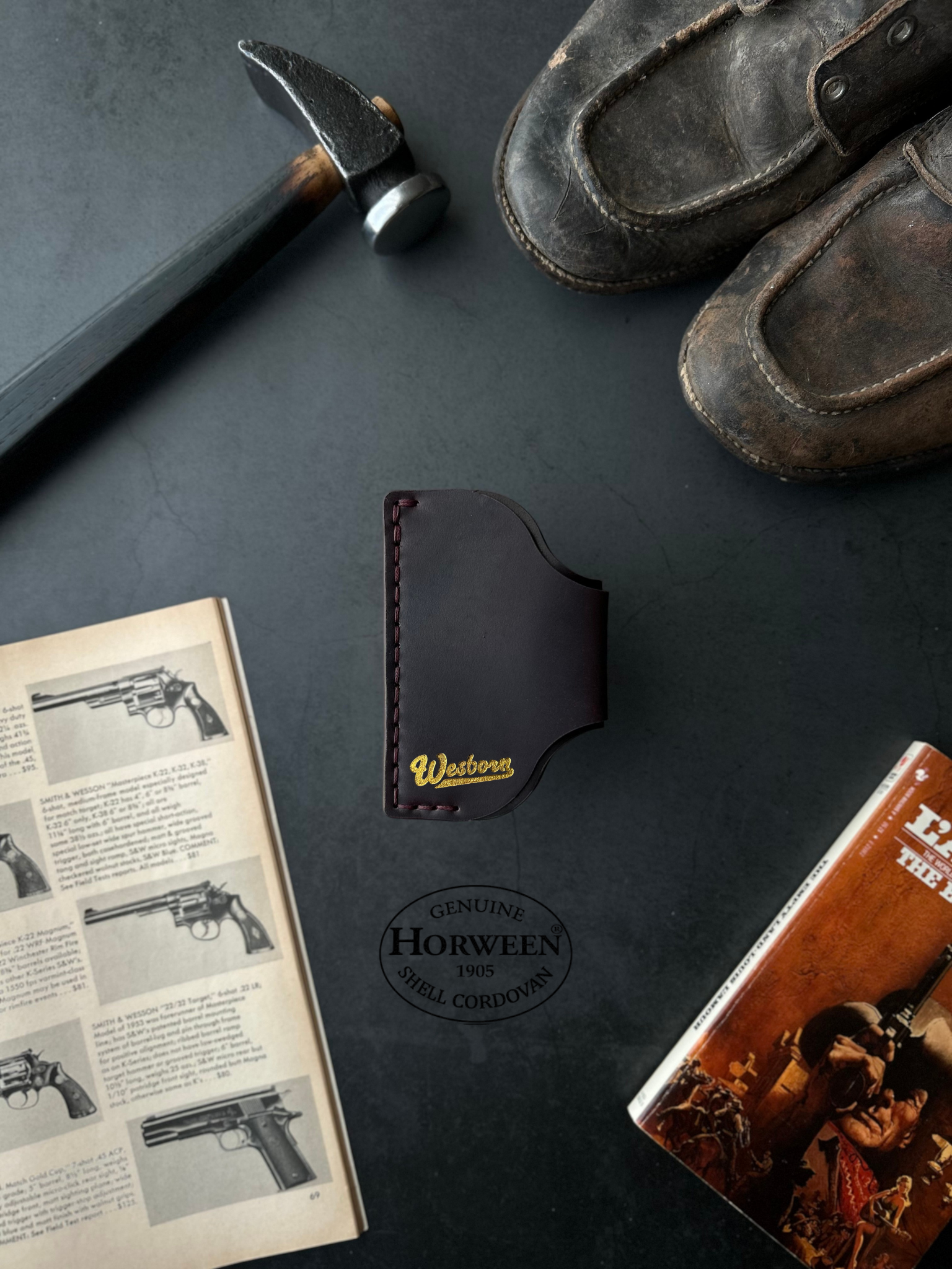 Black leather wallet with gold logo on a dark surface with books and shoes.
