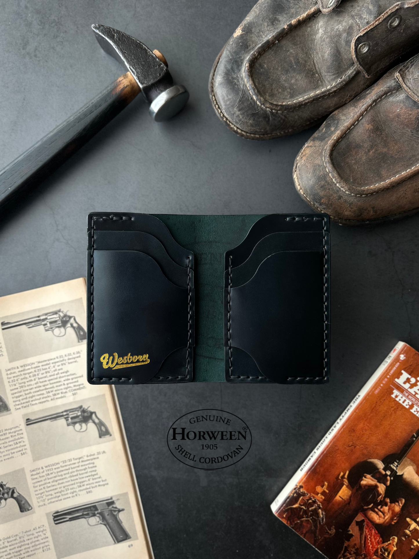 Black leather wallet with gold lettering on a dark surface with shoes, hammer, and book.