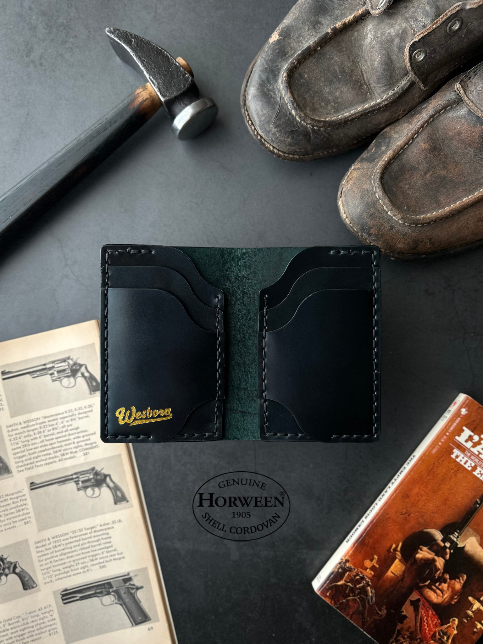 Black leather wallet with gold lettering on a dark surface with shoes, hammer, and book.